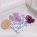 Displayed on a white surface, a charming decorative setup features the "Do Me Now Naughty Candy Heart Purple Butt Plug" by Blush, a heart-shaped purple silicone object with "LOVE NOW" written on it, housed inside a clear round container. Accompanying the display are a wooden lid, a floral-patterned notebook, and red and blue scrunchies. This elegant arrangement is ideal for anal beginners seeking stylish intimacy.