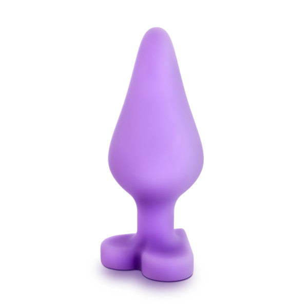 A purple Blush "Do Me Now Naughty Candy Heart" butt plug, featuring a cone shape with a flared base, standing upright against a plain white background. Crafted from soft silicone and designed with ergonomic considerations, this hypoallergenic phthalate-free piece promises both safety and comfort.