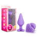 A pink box labeled "Do Me Now Naughty Candy Heart Purple Butt Plug" by Blush contains a purple, heart-shaped butt plug made from soft silicone. Alongside the box, two views of the hypoallergenic, phthalate-free toy are shown: one standing upright and one displaying the words "DO ME NOW" engraved on its heart-shaped base.