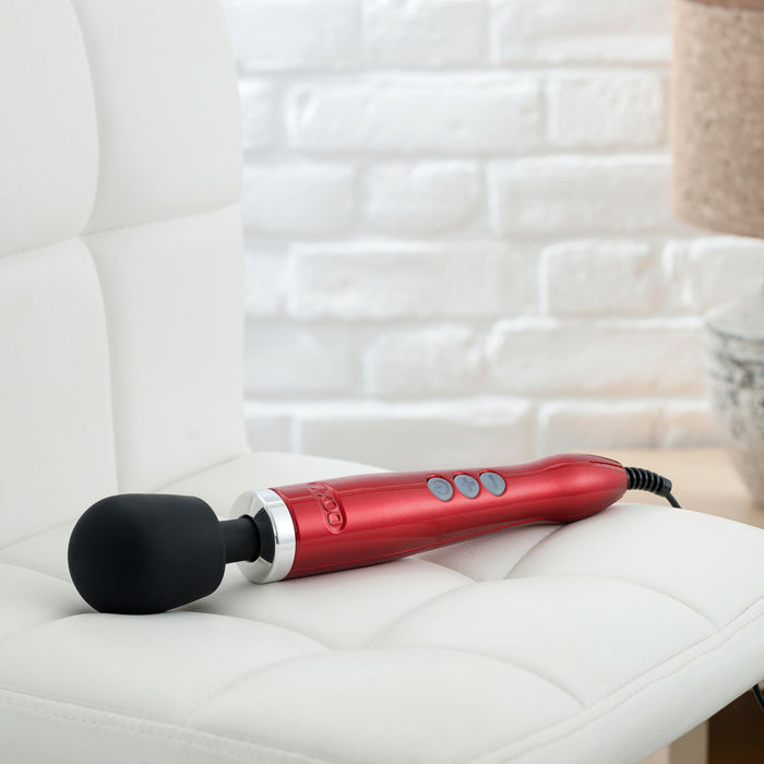 A Doxy Die Cast Extra Powerful Wand Vibrator - Red, with a black cushioned head and deep rumbly vibrations, rests on a white cushioned chair. The red handheld massager features multiple buttons on the handle, set against a background with a partially visible white brick wall.