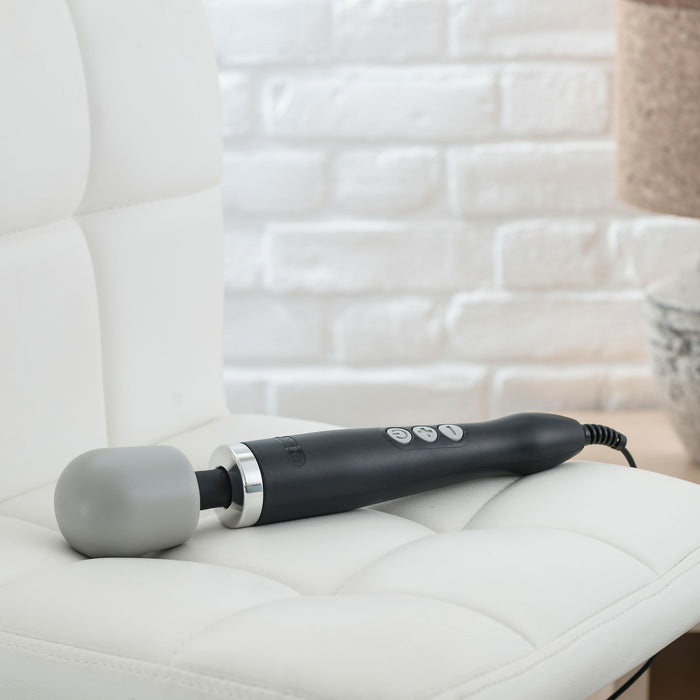 The Doxy Extra Powerful Wand Vibrator - Black, featuring control buttons on its handle and a large, round, gray head, sits on a white cushioned chair. Made from medical-quality silicone and connected to a power cord, the black electric handheld massager stands against a backdrop of a brick wall and part of a lamp.