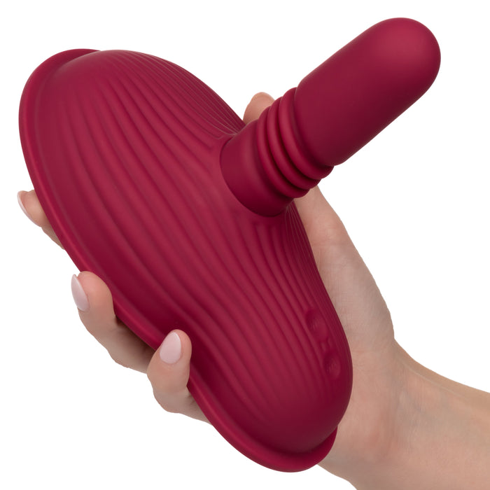 A person's hand holds the Dual Rider Remote Control Thrust & Grind Humping Vibrator from CalExotics. This red, textured sex toy with a rounded base and an extended cylindrical shaft is made of silicone and features ribbed details along with dual motor massager capabilities for enhanced intimate use.