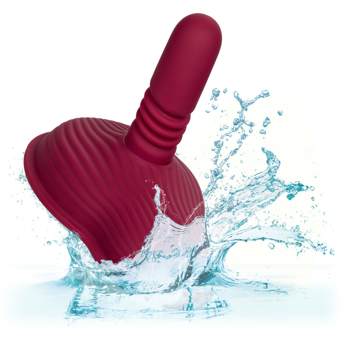 The Dual Rider Remote Control Thrust & Grind Humping Vibrator by CalExotics is depicted partially submerged, creating a splash in clear water. It features a red, ribbed design with a cylindrical handle and a smooth, curved base. The white background accentuates the splashing water around the massager.