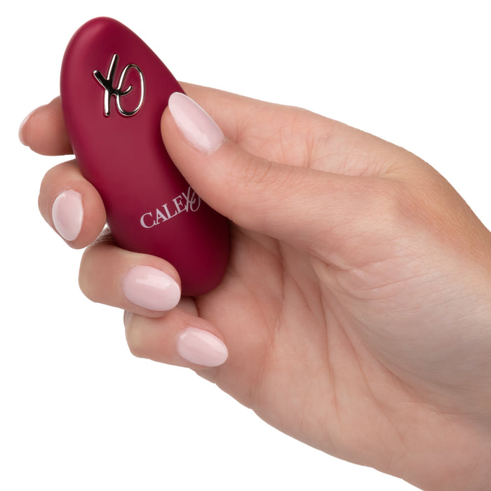 A hand with light pink nail polish holds a small, maroon-colored device. The waterproof massager, branded with "CalExotics" in white letters, is equipped with a metallic switch or button on the top and is identified as the Dual Rider Remote Control Thrust & Grind Humping Vibrator.