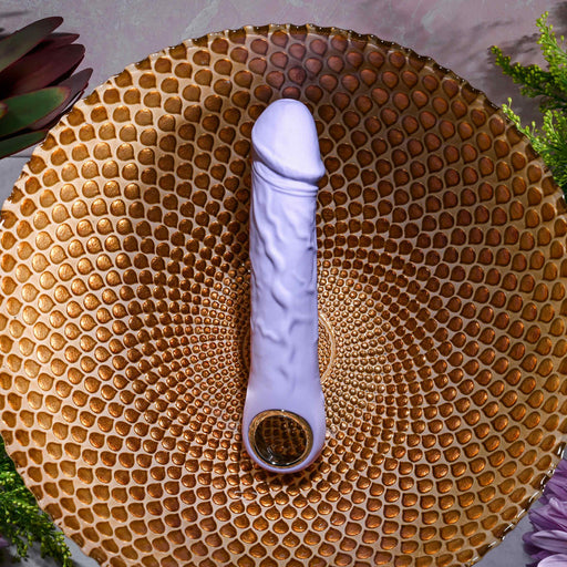 The Purple Fantasy Veiny Silicone Vibrator with Finger Loop, flexible and USB magnetic rechargeable, sits on a textured, honeycomb-patterned golden plate. It's safe silicone body is surrounded by green plants and dark red flowers against a gray background.