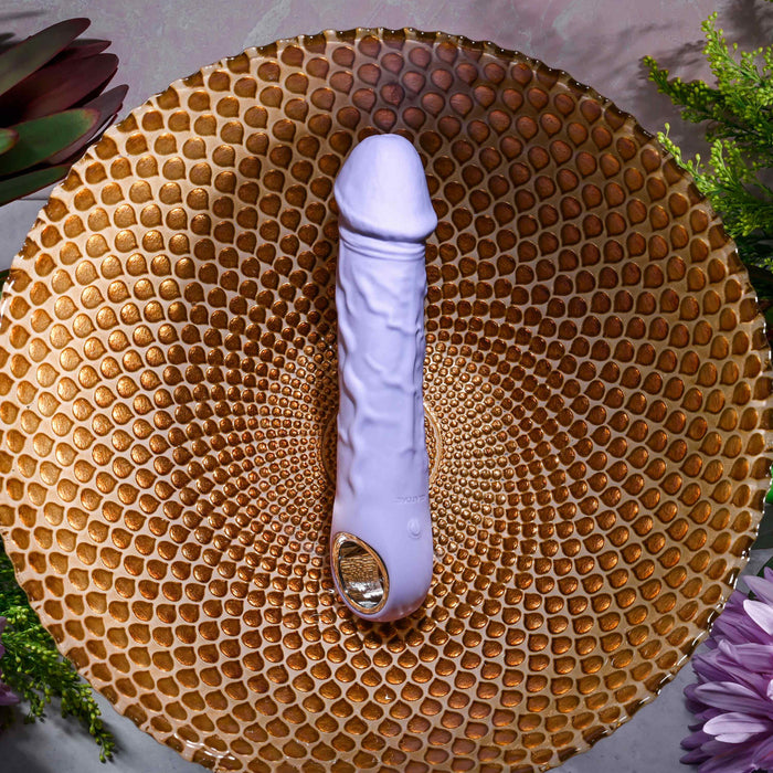 The Purple Fantasy Veiny Silicone Vibrator with Finger Loop, in a soothing lavender hue, sits on a textured gold-toned plate. Graced by green foliage and purple flowers, the setting exudes an inviting and serene vibe.