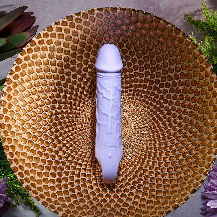 The Purple Fantasy Veiny Silicone Vibrator with Finger Loop lies on a textured, circular gold-patterned surface. The body-safe design is accentuated by green plants and purple flowers, creating a vibrant contrast in the scene.