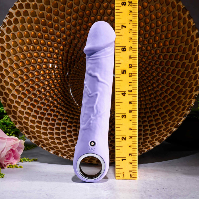 The Purple Fantasy Veiny Silicone Vibrator with Finger Loop, in a vibrant purple hue, stands upright beside a yellow ruler to display its length. A brown decorative bowl sits behind it, with pink flowers and lush greenery framing this body-safe, waterproof toy.