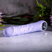 A Purple Fantasy Veiny Silicone Vibrator with Finger Loop, phallic-shaped and light purple, rests on a textured surface. It has detailed texture and a metallic end, surrounded by soft-focus pink flowers and green foliage for a serene setting.