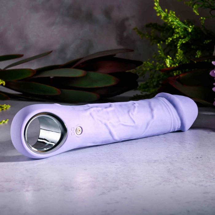 A Purple Fantasy Veiny Silicone Vibrator with Finger Loop sits on stone, surrounded by greenery. It features a sleek, silver handle and body-safe buttons on the side.