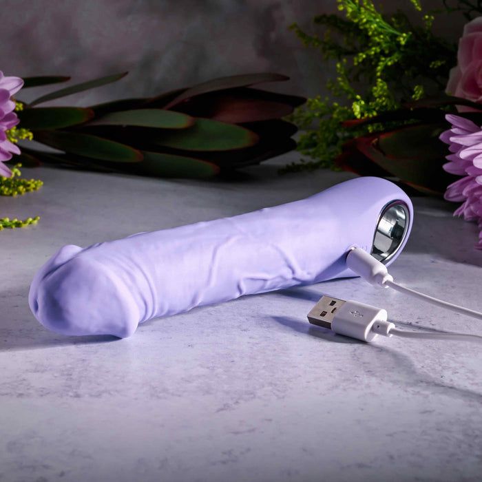 The Purple Fantasy Veiny Silicone Vibrator, shaped like a phallus and featuring a finger loop, rests on marble. It's connected to a USB cable, with blurred pink flowers and green foliage in the background.