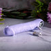 The Purple Fantasy Veiny Silicone Vibrator, shaped like a phallus and featuring a finger loop, rests on marble. It's connected to a USB cable, with blurred pink flowers and green foliage in the background.