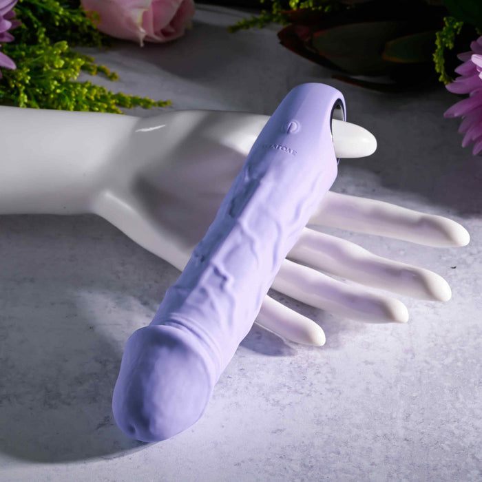 A purple Fantasy Veiny Silicone Vibrator with a finger loop sits on a white mannequin hand against a backdrop of pink flowers and green leaves, enhancing the elegance of this waterproof, body-safe marvel.