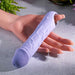 A hand holds the Purple Fantasy Veiny Silicone Vibrator with its lifelike texture and silver finger loop, set against a light surface with greenery. This body-safe device adds elegance and pleasure to any setting.