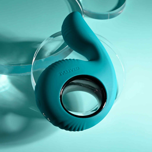 The Thumbs Up Shallow Penetration Vibrator with Finger Loop, in teal silicone, rests on a reflective surface. Its ergonomic, ring-shaped design boasts textured details and adjustable vibrating speeds. A soft teal background enhances its sleek, modern look.