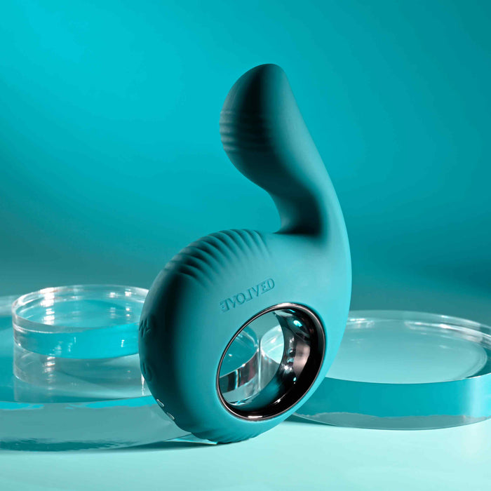 The Thumbs Up Shallow Penetration Vibrator with Finger Loop, teal and ergonomically shaped in silicone with ribbed texture, is elegantly displayed against a turquoise background alongside circular clear acrylic platforms.