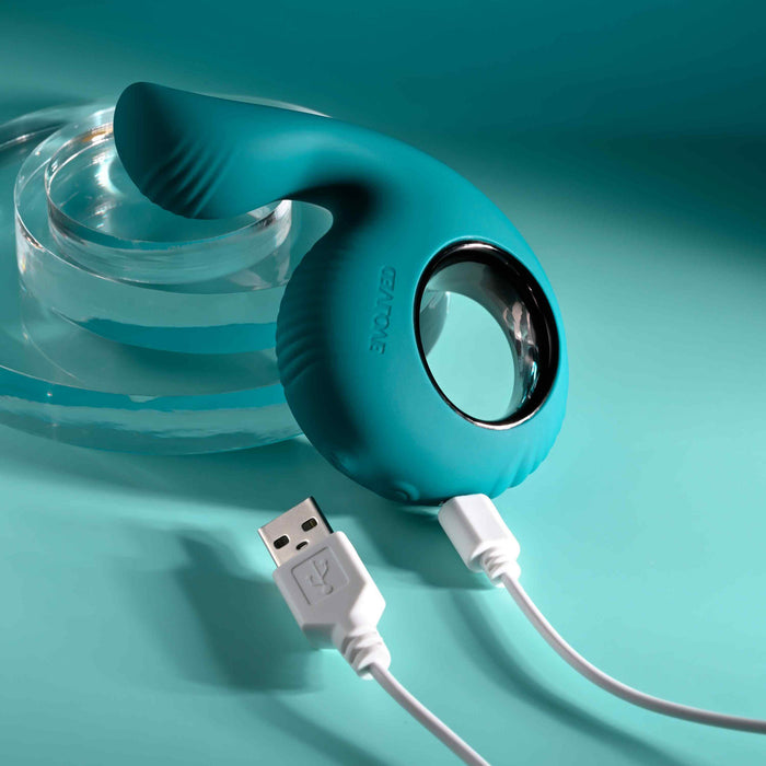 On a turquoise background, the Thumbs Up Shallow Penetration Vibrator with Finger Loop, featuring a ridged teal surface and circular cutout, connects to a coiled white USB charger. A transparent acrylic stand is partially visible in the background, hinting at its potential for powerful anal orgasms.