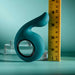 The Thumbs Up Shallow Penetration Vibrator with Finger Loop, a teal silicone toy featuring a textured, curvy design and hole, is displayed alongside a yellow measuring tape on a teal background. Transparent circular platforms at the base enhance its sophisticated allure.