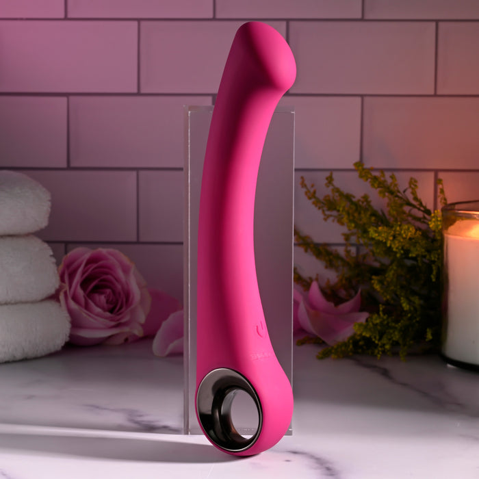 The pink Pleasure Curve G-Spot Vibrator with Finger Loop stands upright on marble amidst neatly folded towels, a pink rose, greenery, and a lit candle. Its waterproof vibrations offer tranquility against the light-colored tiled backdrop.