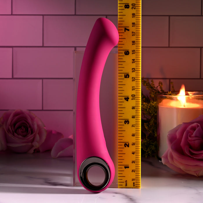 A pink Pleasure Curve G-Spot Vibrator with a silver finger loop stands upright beside a yellow inch measuring tape. In the background, pink roses and a lit candle enhance its allure on the tiled surface.