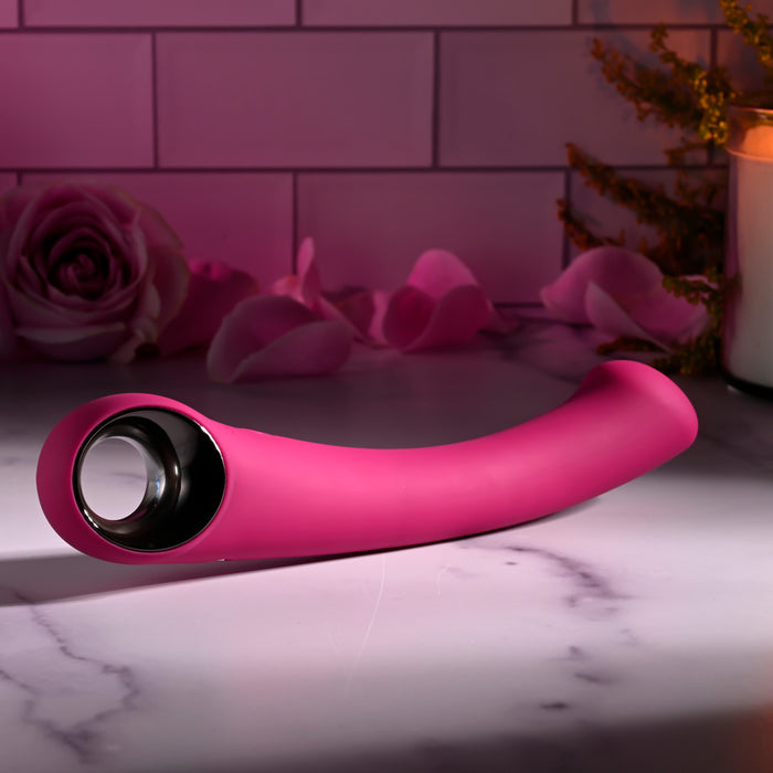 A pink, curved silicone Pleasure Curve G-Spot Vibrator with Finger Loop rests on a marble surface. The scene includes an open rose, petals, a lit candle, and greenery, creating an intimate ambiance enhanced by its soothing waterproof vibrations.