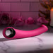 A pink, curved silicone Pleasure Curve G-Spot Vibrator with Finger Loop rests on a marble surface. The scene includes an open rose, petals, a lit candle, and greenery, creating an intimate ambiance enhanced by its soothing waterproof vibrations.