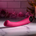 The Pleasure Curve G-Spot Vibrator with Finger Loop, a pink curved electronic device, rests on a marble surface. Soft pink roses and greenery softly glow under a warm-lit candle against white tile walls. It features waterproof vibrations for endless enjoyment.