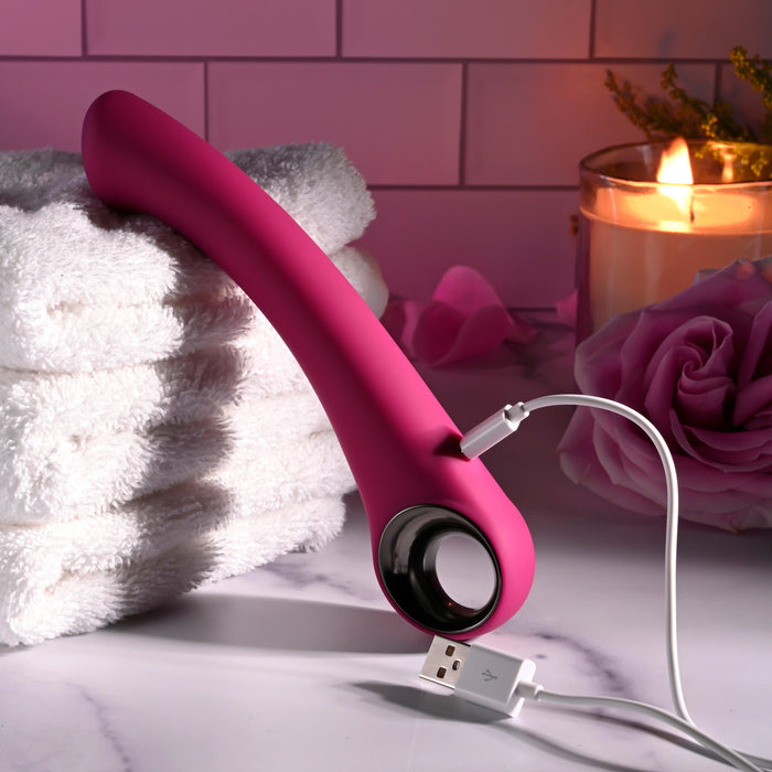 A pink Pleasure Curve G-Spot Vibrator with Finger Loop charges on white towels, creating a serene atmosphere enhanced by a lit candle and pink rose in the background. Its waterproof vibrations contribute to the cozy, relaxed setting.