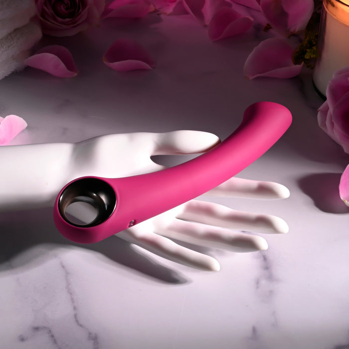 The Pleasure Curve G-Spot Vibrator with Finger Loop in pink is elegantly displayed on a mannequin hand over a marble surface. Surrounded by rose petals and a lit candle, it creates an intimate and softly lit ambiance inviting exploration and relaxation.