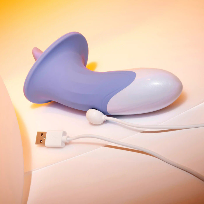 The Lix & Kisses Swirling Tongue Oral Sex Vibrator by Evolved Novelties, featuring a predominantly lavender silicone body with a white detail and a flared base, rests on a light-colored surface. A white USB charging cable is connected to the vibrator, extending towards the foreground. The background showcases a warm gradient lighting effect.