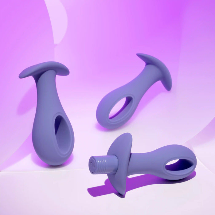 The Hole Punch Vibrating 3 Piece Anal Training Set, featuring three purple silicone plugs with ergonomic designs, is displayed against a vibrant purple background. Each toy has an integrated loop handle for easy grip, varied shapes for diverse uses, and a USB rechargeable system.