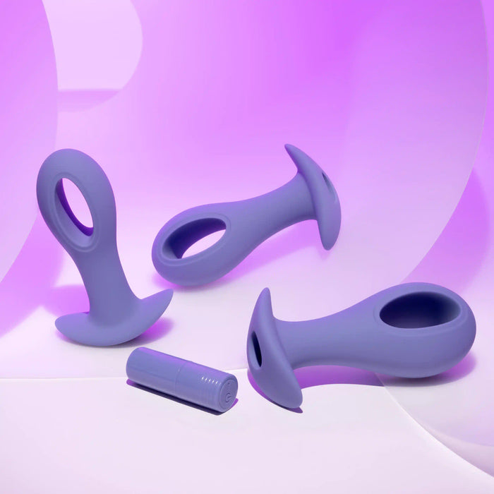 The Hole Punch Vibrating 3 Piece Anal Training Set showcases three purple silicone plugs with flared bases and rounded tips, one of which is split for unique sensations, all set against a soft purple-to-white gradient background.