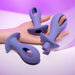 A hand holds the Hole Punch Vibrating 3 Piece Anal Training Set: three purple silicone plugs with unique hollow designs and smooth surfaces, each featuring circular handles. A soft pink and purple background enhances these versatile toys' elegance.