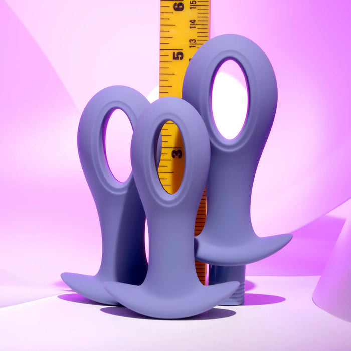 The Hole Punch Vibrating 3 Piece Anal Training Set, featuring purple silicone plugs with round handles and a sleek, hollow design, is shown in front of a yellow measuring tape against a pink-to-purple gradient background.