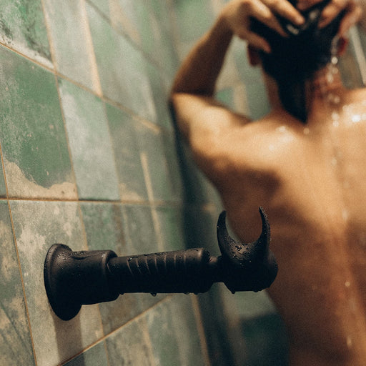 A person is showering, facing away from the camera, with water cascading down their back against the green tiled wall. A unique wrench-shaped black handle subtly enhances the scene, resembling an Evo Vibrating Male Stroker and Couples Vibrator for a touch of intense vibration.