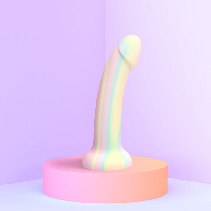 A pastel-colored, phallic-shaped object stands upright on a pastel pink circular platform against a soft purple background. This pegging dildo, the Fantasia Glow in the Dark 7 inch Silicone Dildo by Lovely Planet, features smooth contours and a subtle gradient of soothing colors.