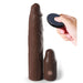 A hand holding a remote control near the Fantasy 9 Inch Vibrating Silicone Penis Extension with Remote Control in Chocolate by Pipedream Products. The penis extender stands upright next to a shorter, rounded device featuring a button on its front and offering 10 modes of vibration. Both objects appear to be made of similar material.