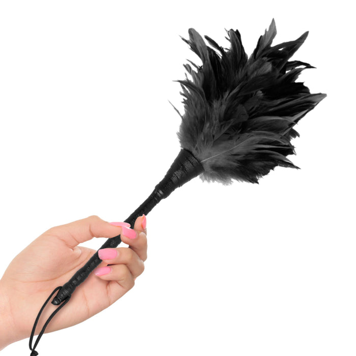 A person's hand with pink nail polish elegantly holds the Fetish Fantasy Frisky Feather Duster by Pipedream Products against a white background. The duster features a black handle and soft, fluffy black feathers at the end, perfect for delivering a sexy stroke.