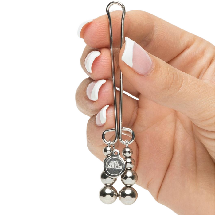 A hand with manicured nails holding a Fifty Shades of Grey Just Sensation Beaded Clitoral Clamp, featuring three attached metal beads and reminiscent of refined elegance. A logo featuring the word “DALLE” is imprinted on a circular part near the beads. The background is white.