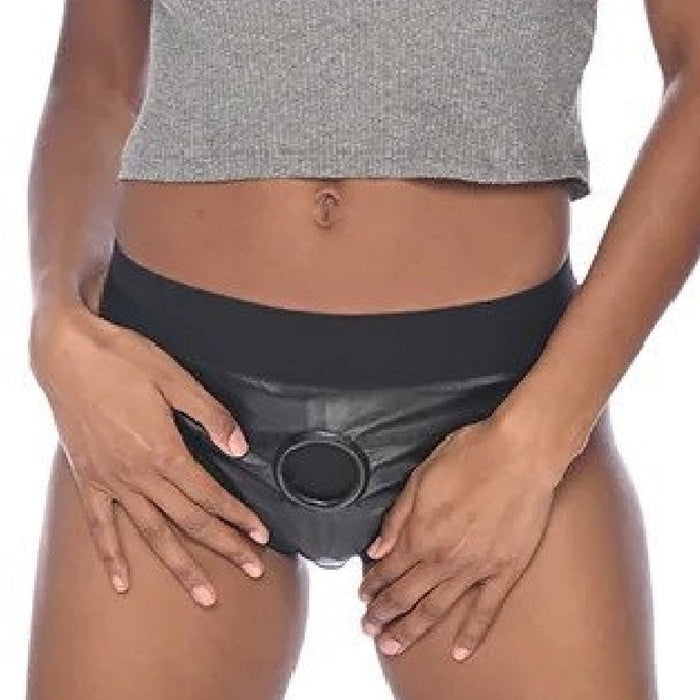 A person dressed in a gray crop top and black leather high-waisted bottoms with an O-ring detail stands with their hands resting on their hips. The bottoms feature the Sportsheets Fit Patent Leather Look Corset Back Strap-on Harness with 2 Bullet Pockets, accentuating the exposed midriff.