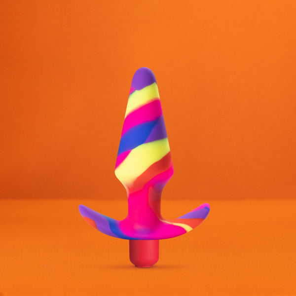 A striking, cone-shaped silicone object with a flared base, showcasing a lively swirl of pink, blue, yellow, and purple hues, is displayed against an orange background. Named the "Free Spirit Vibrating 10 Function Beginner Silicone Butt Plug" by Blush, this sex toy is made from platinum-cured silicone and stands upright, making it perfect for beginners.