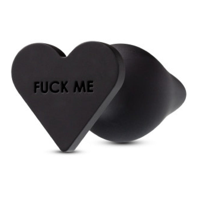 A black, heart-shaped Fuck Me Silicone Heart Butt Plug by Blush, featuring the phrase "FUCK ME" engraved on it. Made of smooth and flexible silicone material, it has a heart-shaped base.