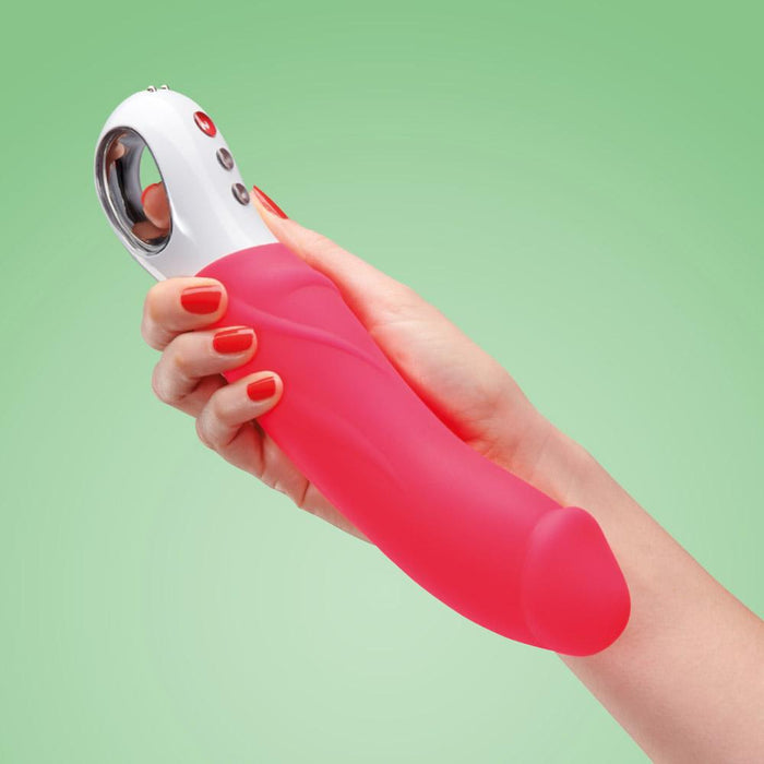 A hand with red-painted nails holds a pink and white Fun Factory Big Boss Thick Vibrator against a green background. The USB rechargeable vibrator from Fun Factory features a curved handle with buttons and a metallic ring at the base, perfect for g-spot stimulation.