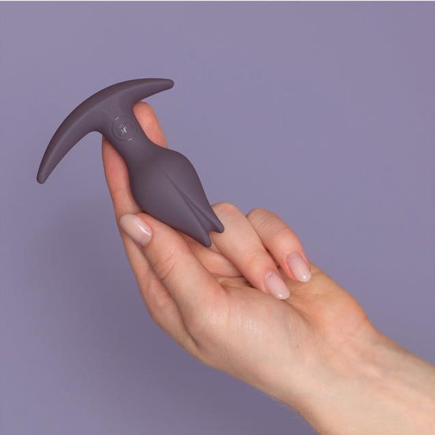 A hand with manicured nails holds a small, sleek, purple silicone device against a light purple background. The Fun Factory Bootie Fem Anal Plug features an ergonomic design with a curved, elongated tip and two side extensions, resembling a minimalist abstract form.