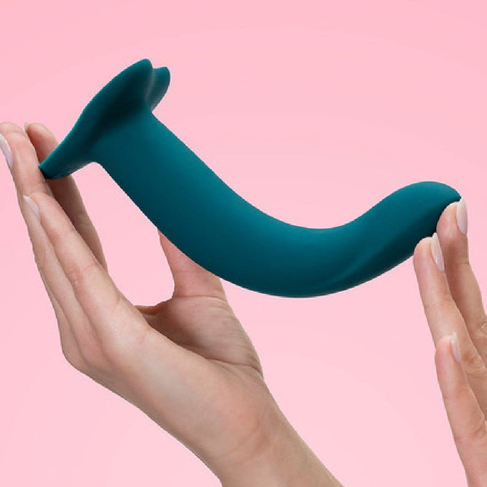 A person's hand holding a Fun Factory Limba Flex Medium Silicone Dildo in Deep Sea Blue with a suction base against a pink background. The device appears sleek and modern in design.