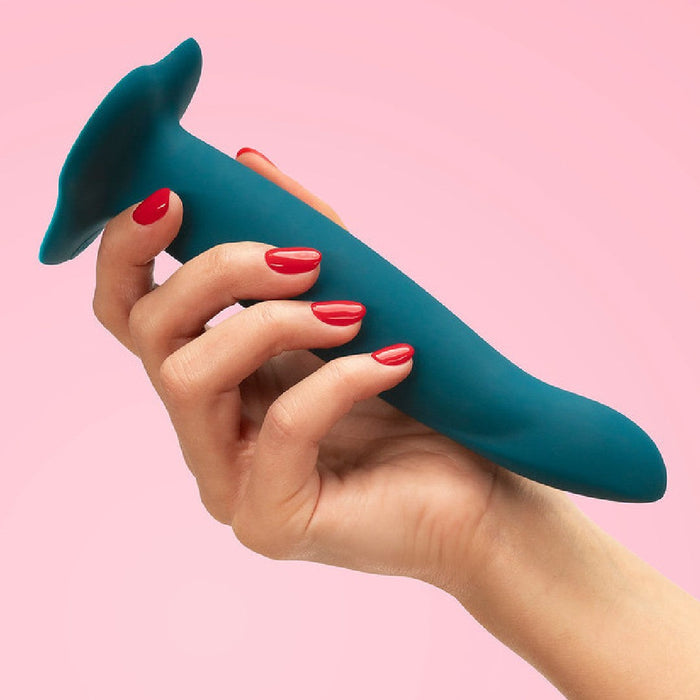 A hand with red nail polish holding a Fun Factory Limba Flex Medium Silicone Dildo - Deep Sea Blue against a pink background.