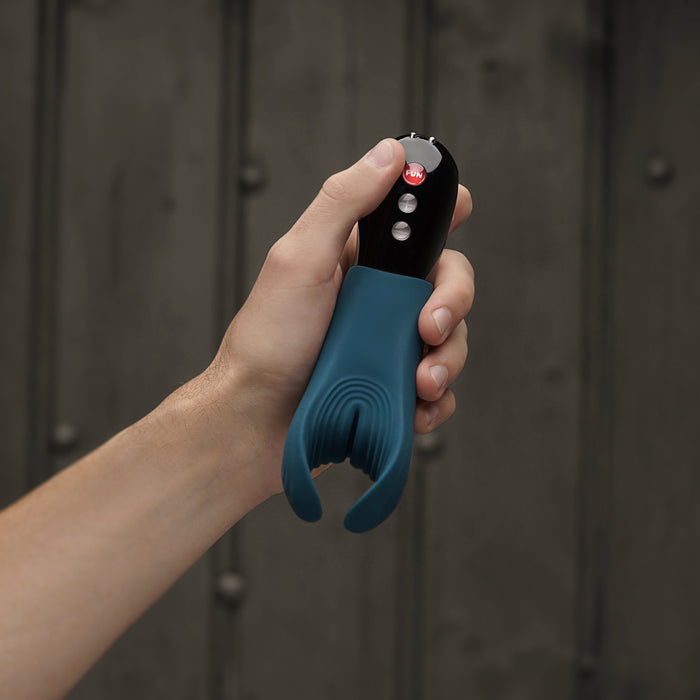 A hand holds a Fun Factory Manta Vibrating Penis Stroker in Deep Blue, featuring a curved, split end design crafted from body-safe silicone. This device includes three buttons on the handle, and the background is an out-of-focus dark surface.