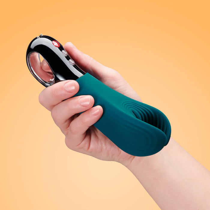 A person holding the Fun Factory Manta Vibrating Penis Stroker - Deep Blue, a sleek, hand-held electronic device from Fun Factory with a chrome and black handle featuring several buttons. Made from body-safe silicone, it has a rounded, split tip and is set against a gradient orange background.