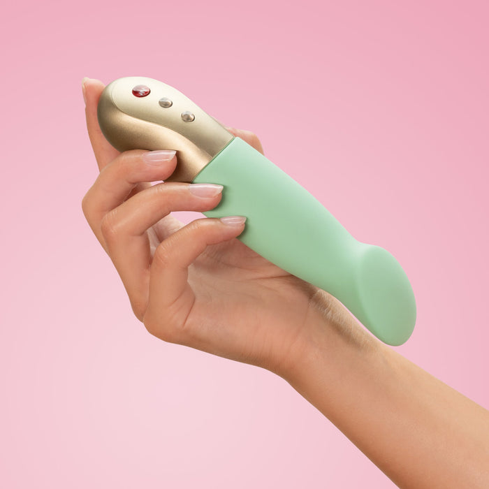 A hand holding a pistachio and gold Fun Factory Sundaze Thrusting Vibrator with buttons. The background is a soft pink color.