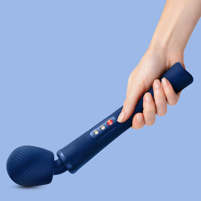 A close-up image of a hand holding the Fun Factory Vim Silicone Weighted Rumbly Wand Vibrator in blue against a light blue background. The powerful wand, from Fun Factory, features a rounded, ridged head, a textured grip, and several control buttons on the handle. Experience deep vibrations for ultimate relaxation.